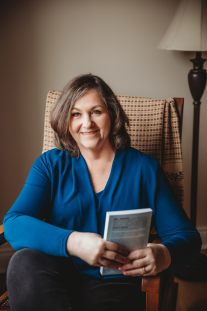 brunette haired woman smiling wearing blue cardigan holding book