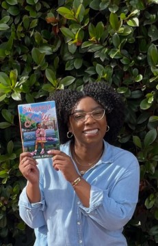 Black Panamanian debut author Tamika Burgess holds her debut novel. She is smiling, has a blue shirt on, has curly dark hair, and wears glasses.